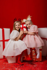 two little girls in elegant white dresses on a red background in a studio with gift boxes catching confetti for the New Year