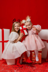 two little girls in elegant white dresses on a red background in a studio with gift boxes catching confetti for the New Year