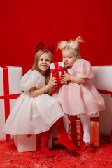 two little girls in elegant white dresses on a red background in a studio with gift boxes catching confetti for the New Year
