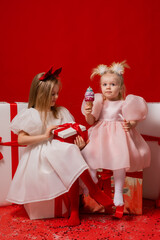 two little girls in elegant white dresses on a red background in a studio with gift boxes catching confetti for the New Year