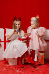 two little girls in elegant white dresses on a red background in a studio with gift boxes catching confetti for the New Year