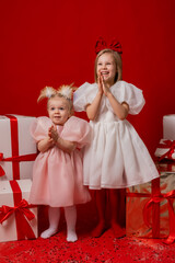 two little girls in elegant white dresses on a red background in a studio with gift boxes catching confetti for the New Year