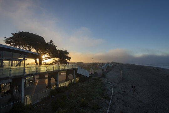 View of the sun kissing the horizon, casting a golden glow on the beachside building and the misty shoreline, a tranquil scene, Christchurch, Canterbury Region, New Zealand.
