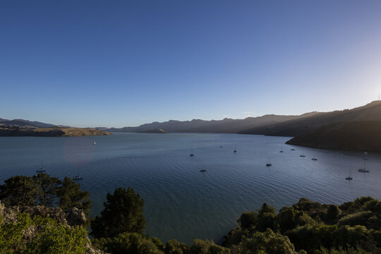View of the sun kissing the tranquil waters where sailboats dance gently, framed by verdant hills under a vast, clear sky, Christchurch, Canterbury Region, New Zealand.