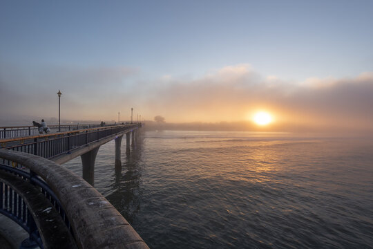 View of the pier stretches into the misty horizon, reflecting the golden sunrise over the calm waters, a peaceful scene of early morning, Christchurch, Canterbury Region, New Zealand.