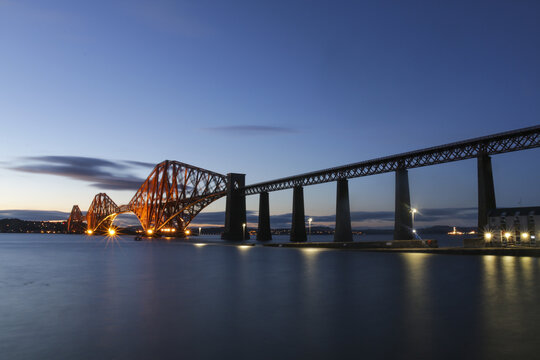 View of the illuminated Forth Bridge stretches across the calm, dark waters under a twilight sky, a beacon of engineering marvel, Edinburgh, Scotland, United Kingdom.