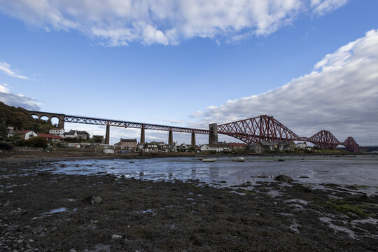 View of the iconic red Forth Rail Bridge stretches across the Firth of Forth under a bright sky, contrasting with the tidal landscape, Edinburgh, Scotland, United Kingdom.