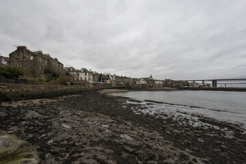 Edinburgh, United Kingdom - 07 December 2016: View of the Firth of Forth's muddy shoreline, a stone wall, and quaint buildings beneath a grey sky, with the majestic Forth Bridge spanning the horizon.