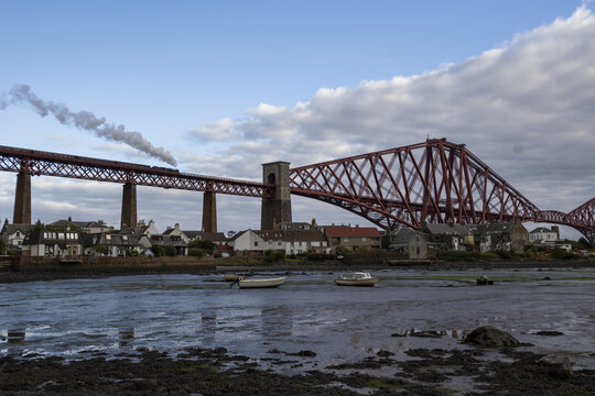 View of a red railway bridge stretches across the water, a train puffing smoke as it crosses above a quaint village, Edinburgh, Scotland, United Kingdom.