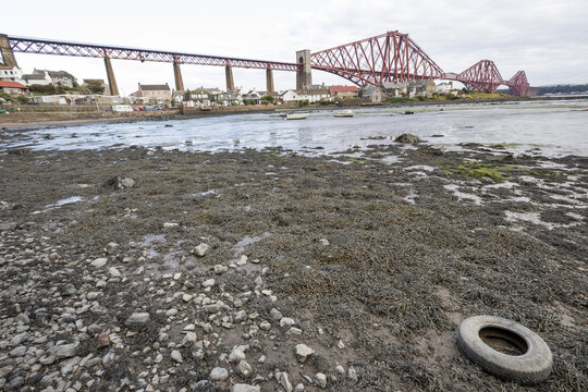 View of the iconic red Forth Bridge arches over the Firth of Forth, contrasting against the muddy shore and cloudy sky, Edinburgh, Scotland, United Kingdom.