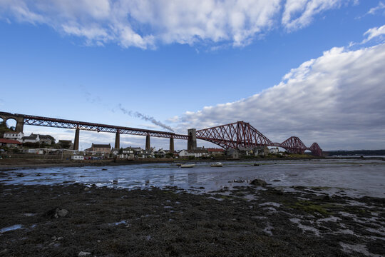 View of the iconic red Forth Bridge piercing the horizon, connecting lands across the glistening Firth of Forth, under a bright, cloud-streaked sky, Edinburgh, Scotland, United Kingdom.