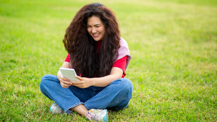 Joyful Asian woman sitting cross-legged on grass reading an electronic book in park