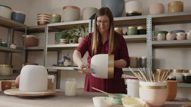Woman painting a handmade clay pot in a ceramics workshop. Artisan applying green paint with a brush, crafting and decorating pottery in an art studio