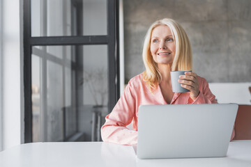 Smiling mature woman sit at desk at home work online on computer drink coffee tea from mug. Happy aged female businesswoman enjoy morning in cozy living room use laptop for distant job.