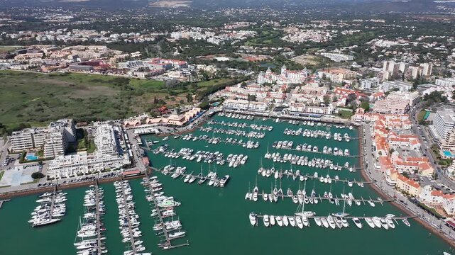 Vilamoura aerial view Aerial view Vilamoura marina Algarve Portugal, sunlit turquoise harbor with rows of moored yachts, busy docks and waterfront promenade, resort buildings and green hills
