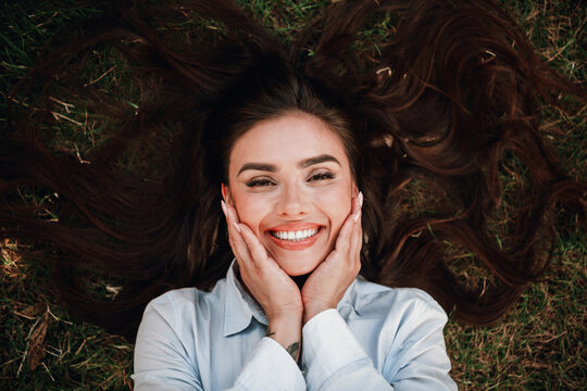 Playful sunny selfie of a stylish young woman lying on grass and smiling with hands on her cheeks in a joyful fashion photo