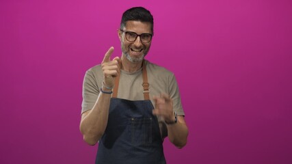 Man pointing finger while smiling and wearing denim apron and glasses in studio with magenta background; friendly welcome.
