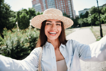 laughing stylish woman selfie in sunny park wearing straw hat light blue shirt and beige top with urban spring vibe and carefree fashion moment
