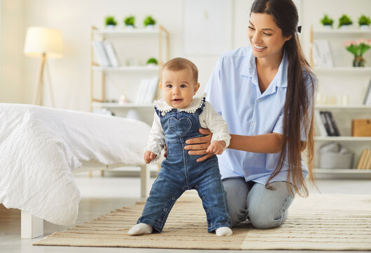 Portrait of happy cheerful mother sitting with her infant little cute child daughter supporting her in first steps at home. Toddler girl stepping on floor with mom help. Family and care concept. - Powered by Adobe