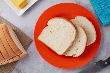 Three (3) slices of Jamaican hard dough bread on an orange plate with a bread knife, stick of butter and a kitchen towel