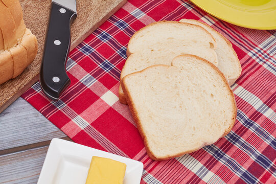 Three slices of Jamaican hard dough bread on a Jamaican bandana with a yellow plate, cutting board, knife and a stick of butter