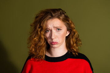 Young woman with red curly hair wearing a red and black sweater looks puzzled against an olive background