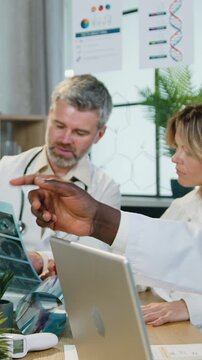 Good-looking successful purposeful experienced multiethnic male and female medical workers sitting around workplace in modern clinic office and discussing x-ray image and get aquainted with different