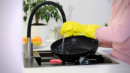 Woman in bright yellow rubber gloves meticulously washing a black frying pan under running water from the faucet in a modern kitchen sink, embodying a daily household chore and hygiene