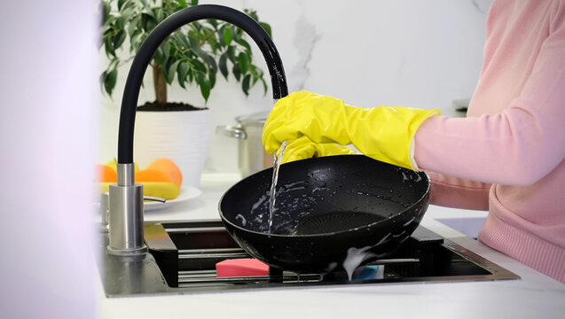 Woman in bright yellow rubber gloves meticulously washing a black frying pan under running water from the faucet in a modern kitchen sink, embodying a daily household chore and hygiene