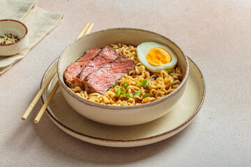 Bowl filled with ramen noodles, tender sliced beef, boiled egg and fresh green onions, asian style soup on light beige background