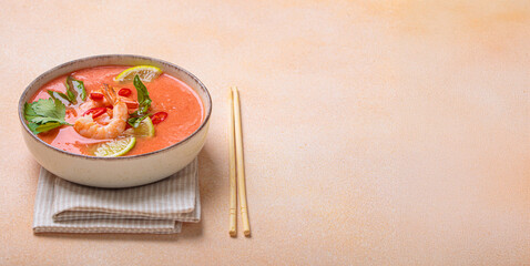 Creamy flavourful tom yum soup, a popular dish of Thailand, is served in a bowl, garnished with fresh shrimp, lime wedges, herbs, and chili peppers, accompanied by chopsticks on a linen napkin, copy
