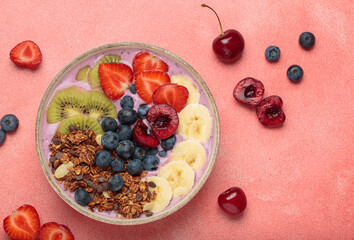 Vibrant and nutritious acai bowl topped with sliced kiwi, strawberries, blueberries, cherries, banana, and granola, a colorful and healthy meal or breakfast on a pink background