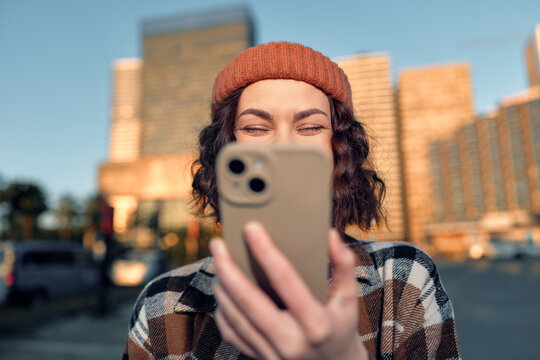 woman with smartphone in city, smiling with beanie and warm sunlight, candid lifestyle portrait capturing authenticity, golden hour glow, mindful living and emotional storytelling in modern urban
