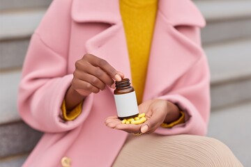 Close-up of a woman in a soft pink trench coat holding a bottle of vitamins, with yellow pills in her hand, showcasing health and wellness lifestyle choices