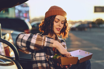 Woman with takeaway food by car trunk in outdoor evening light, casual beanie style and authentic...