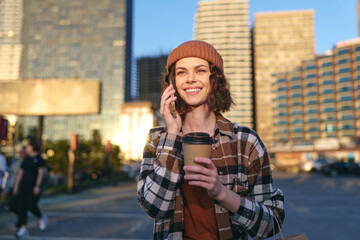 Fototapeta premium woman holding coffee and phone in urban street, smile in golden hour glow, candid lifestyle portrait emphasizing authenticity, mindful living and emotional storytelling in city scene