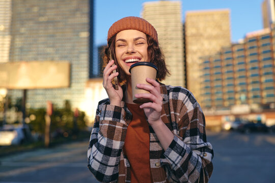 woman coffee phone smile urban beanie plaid walking, candid lifestyle portrait of a young woman holding a takeaway cup and talking on mobile with authenticity, golden hour glow, mindful living
