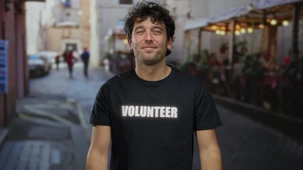 Handsome hispanic man with volunteer shirt smiling and giving thumbs up on a street with an outdoor cafe in the background.