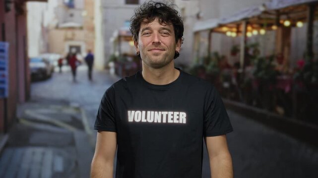 Handsome hispanic man with volunteer shirt smiling and giving thumbs up on a street with an outdoor cafe in the background. - Powered by Adobe