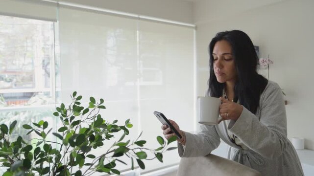 Medium shot of an adult woman in a robe sitting by a bright window, checking her smartphone while holding a coffee mug. 