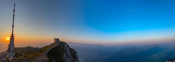 Panoramic sunrise illuminates the transmission tower and chapel on the summit of Dobratsch mountain, revealing vast, hazy alpine landscape and distant Julian Alps under clear sky with a crescent moon