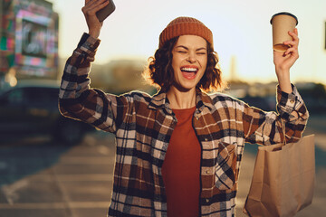Woman with beanie holding coffee and takeaway bag, smiling with phone in city morning, shopping...