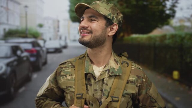 Young hispanic man in camouflage uniform stands smiling with dog tags visible on a street lined with parked cars and leafy hedges; pride.