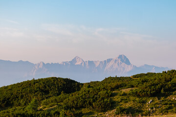 The majestic peaks of the Julian Alps, including Triglav, rise above the horizon, viewed from the lush green alpine meadows of Dobratsch in Carinthia, Austria, under a soft pastel sunrise sky.