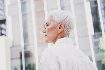 Elegant senior woman walking in the city outdoors wearing a white shirt and glasses expressing confidence and professionalism amid modern office towers in daylight
