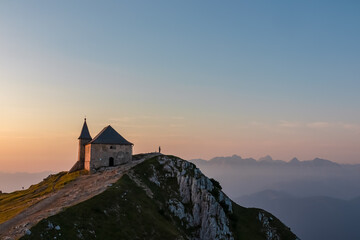 Lone hiker stands in awe on rocky ridge beside the Maria am Stein chapel on Dobratsch, watching spectacular sunrise over fog-filled Julian Alps, embracing a moment of solitude and freedom in Austria.