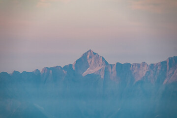 Iconic peak of Mount Triglav is illuminated by gentle first light of sunrise, majestically rising above layers of blue morning mist in the Julian Alps, as seen from Dobratsch in Carinthia, Austria.