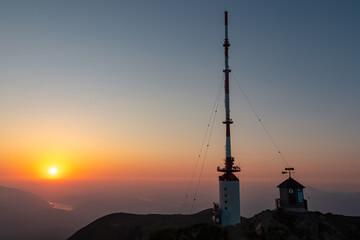 Prominent transmission tower and weather station dominate summit of Dobratsch, silhouetted against magnificent sunrise with sun glowing above a river valley and hazy mountains in Carinthia, Austria.
