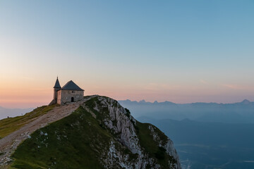 The historic church Maria am Stein stands proudly on the summit of Dobratsch in Carinthia, Austria, with a winding path leading to it against a spectacular panoramic backdrop of the Alps at sunrise.
