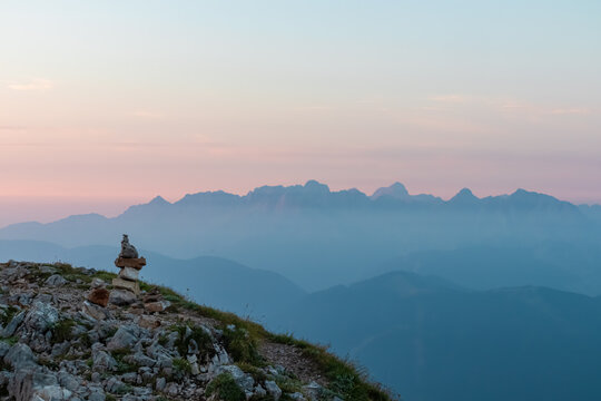 Stone cairn marks a hiking trail on a rugged mountaintop, guiding the way towards a magnificent panoramic view of distant, mist-covered mountain ranges under a serene and soft-colored pastel dawn sky.